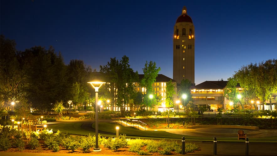A nighttime shot of Green Library with lights shining in the windows, and Hoover Tower in the background.