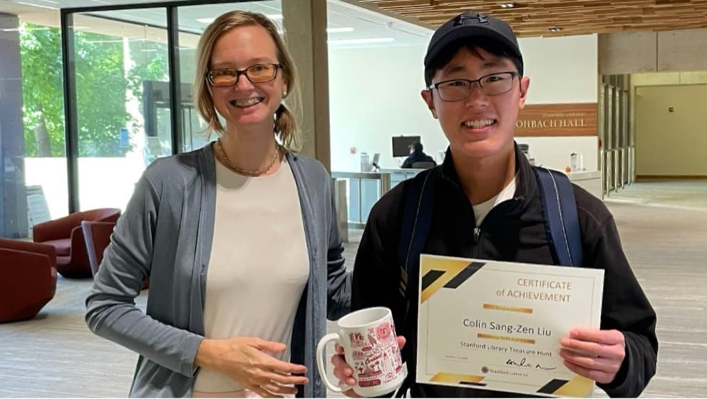 Woman and man standing next to each other, smiling. Man is holding a Certificate of Achievement and mug.