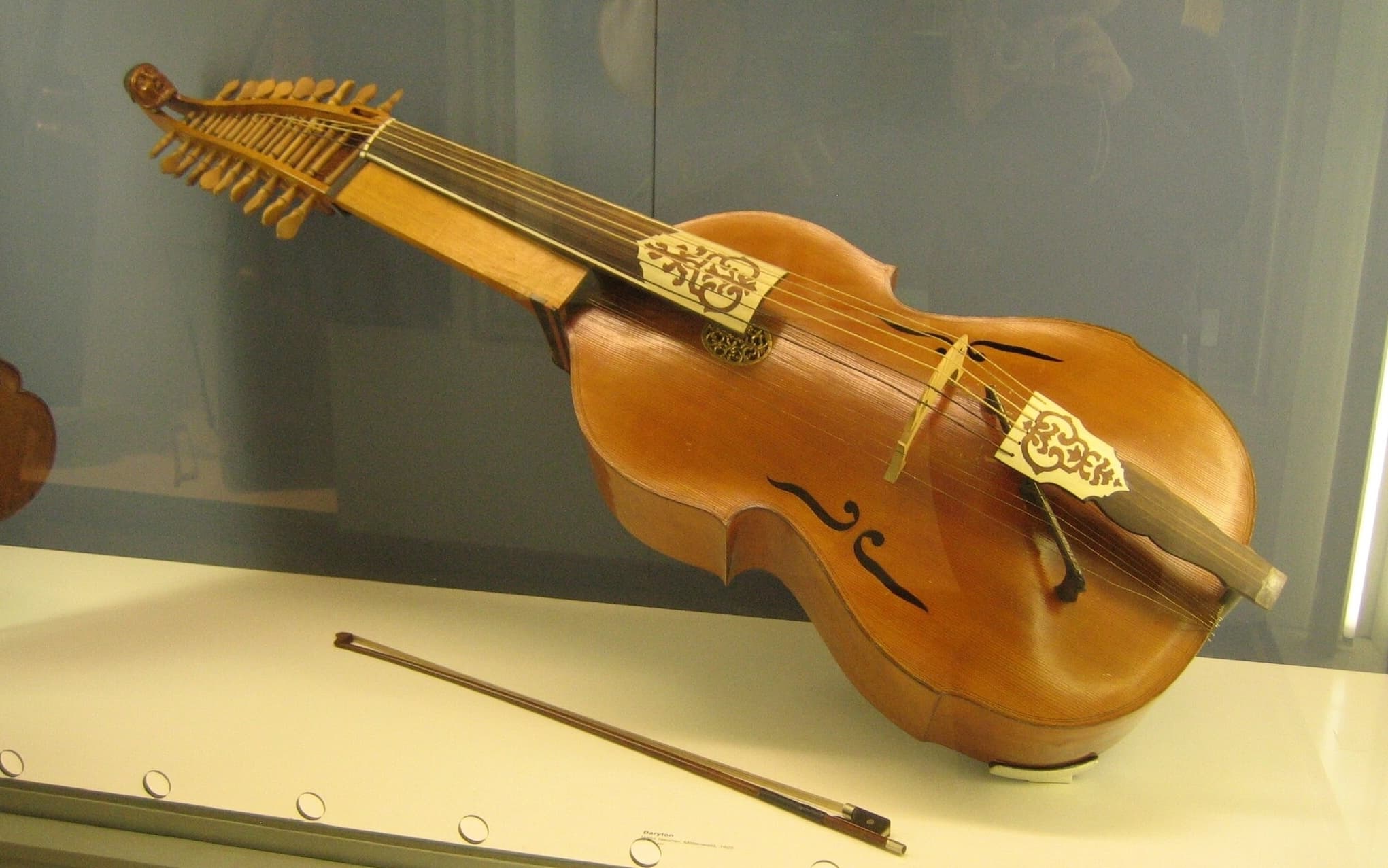 Baryton in display case, showing six bowable strings and twelve sympathetic strings beneath them.  Also shown is a bow.