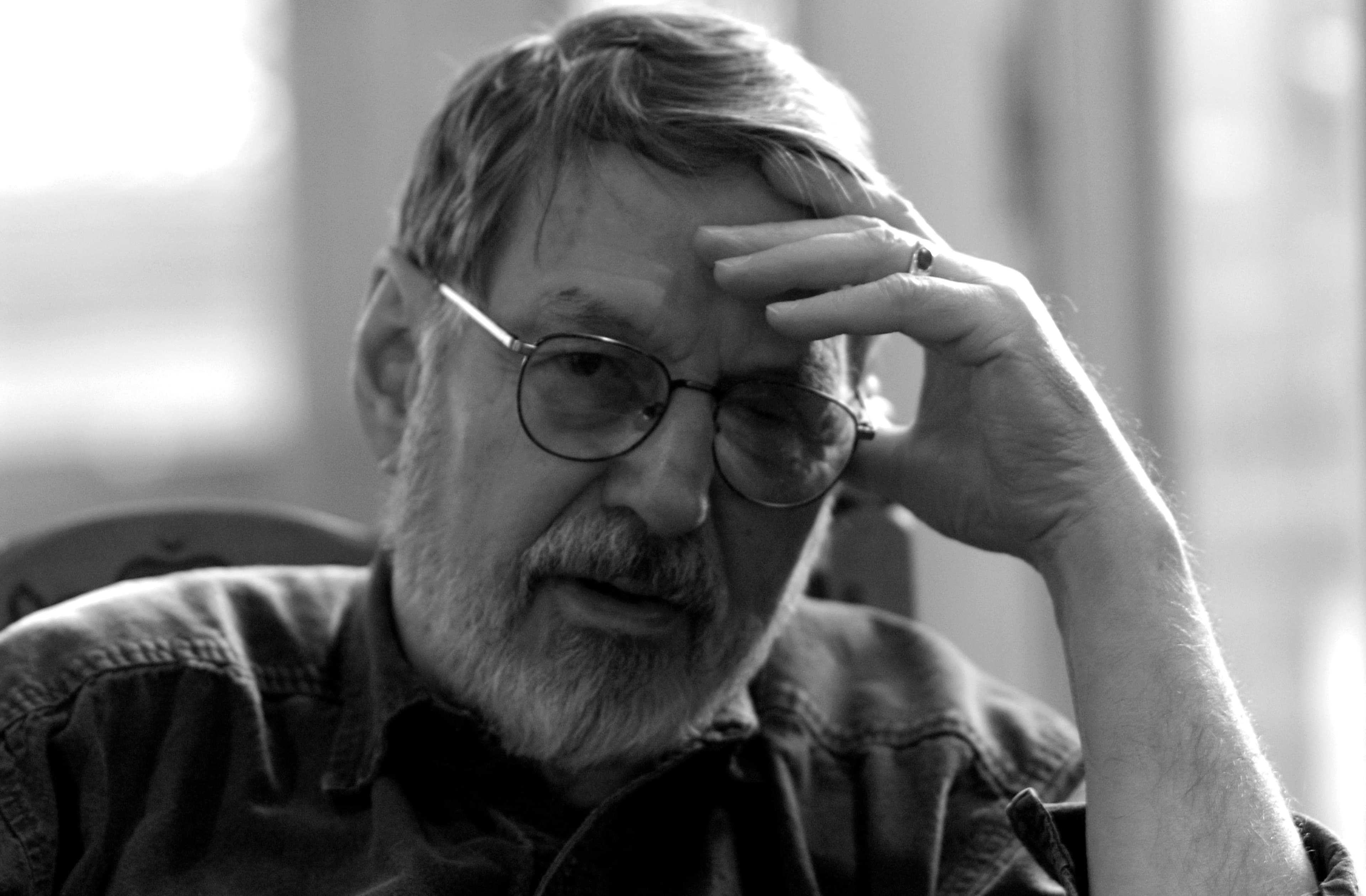 Black and white image of Robert Creeley wearing glasses with left hand on his forehead
